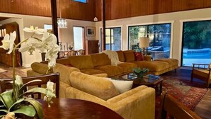 A spacious living room with a large sofa set, a coffee table, and a dining table. There are large windows offering a view of the outdoors. - Mid-mod, boho home w/ a pool! (St. Augustine)