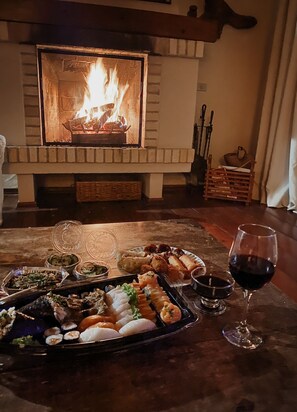 A dining table with a variety of sushi and sashimi, a glass of red wine, and a fireplace with a fire. - Country Home (Campos do Jordão)