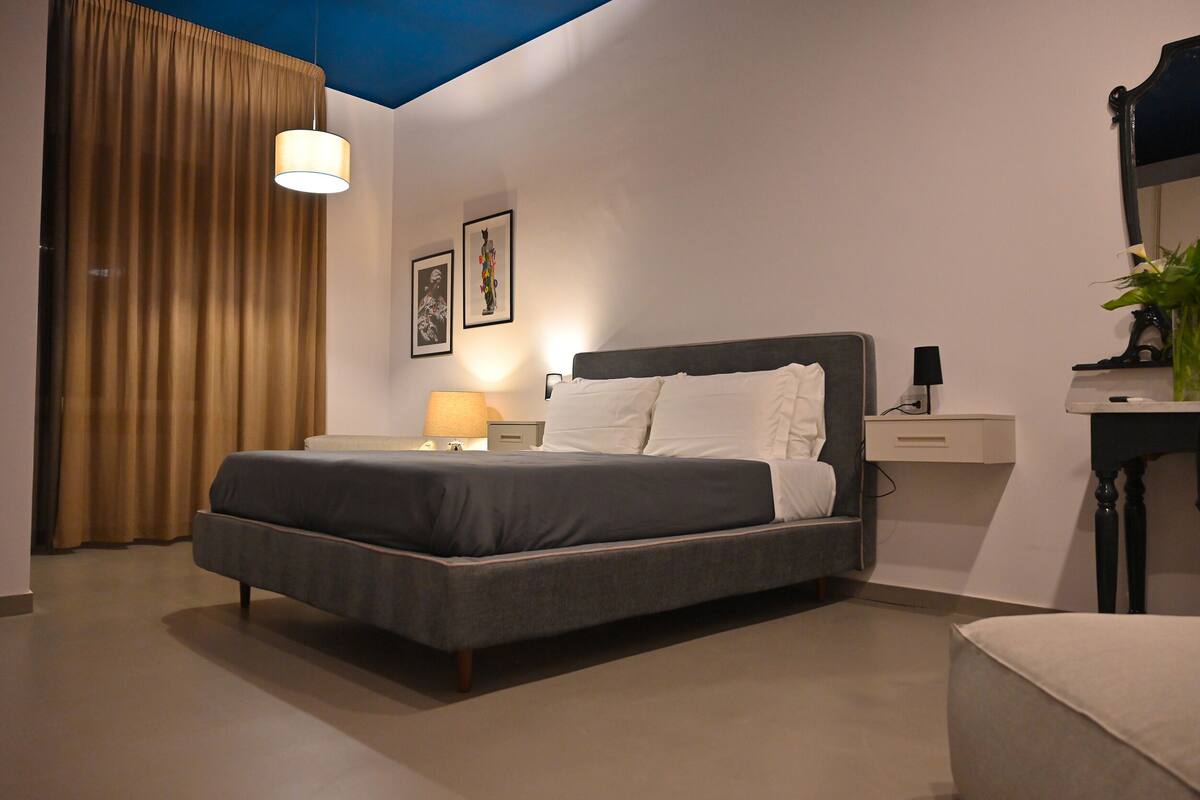 Ein modernes Schlafzimmer mit einem großen Bett, einem Nachttisch, einer Lampe, einem Spiegel und einem Vorhang.