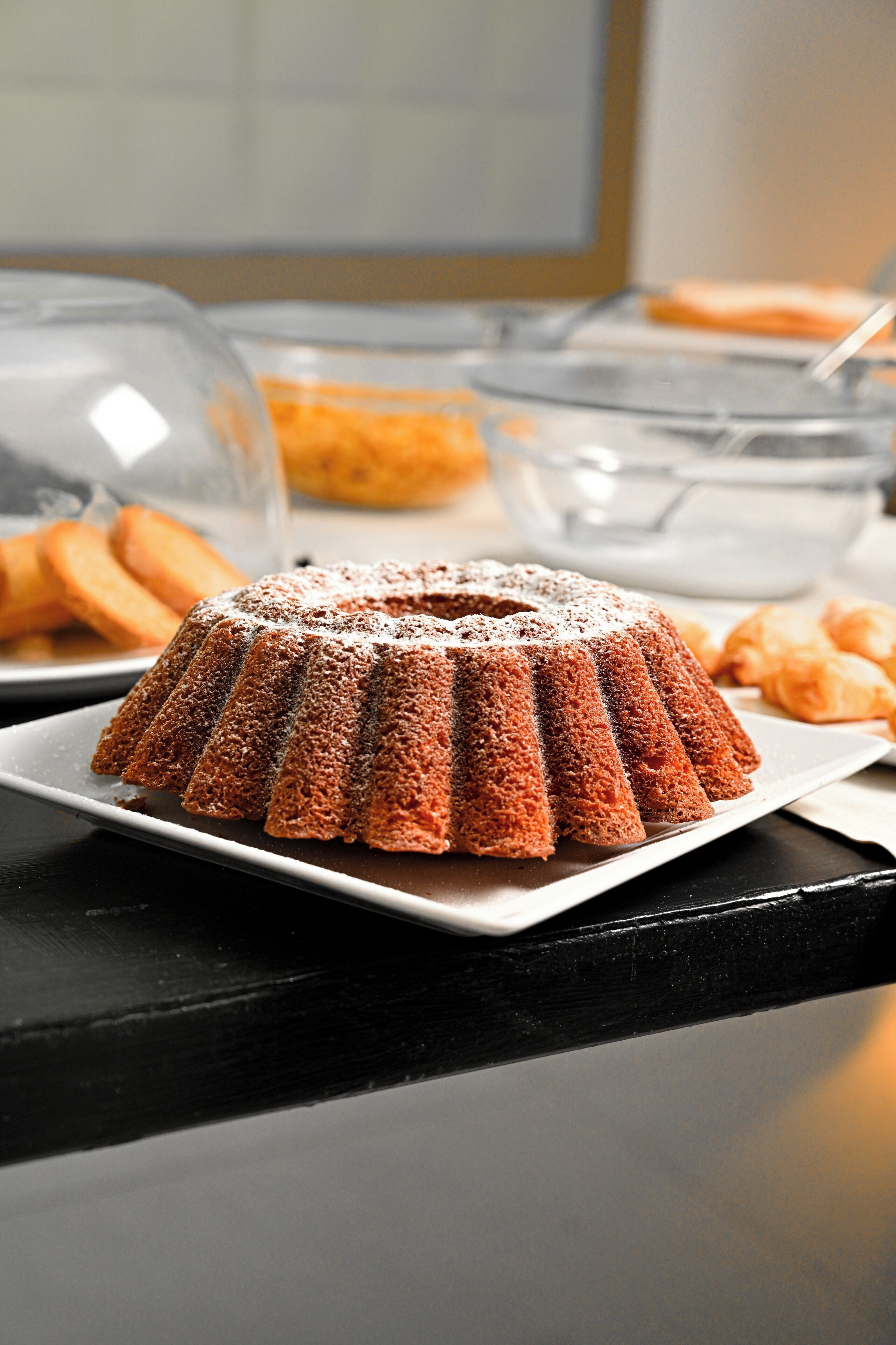 A bundt cake on a plate with a clear glass cover in the background.