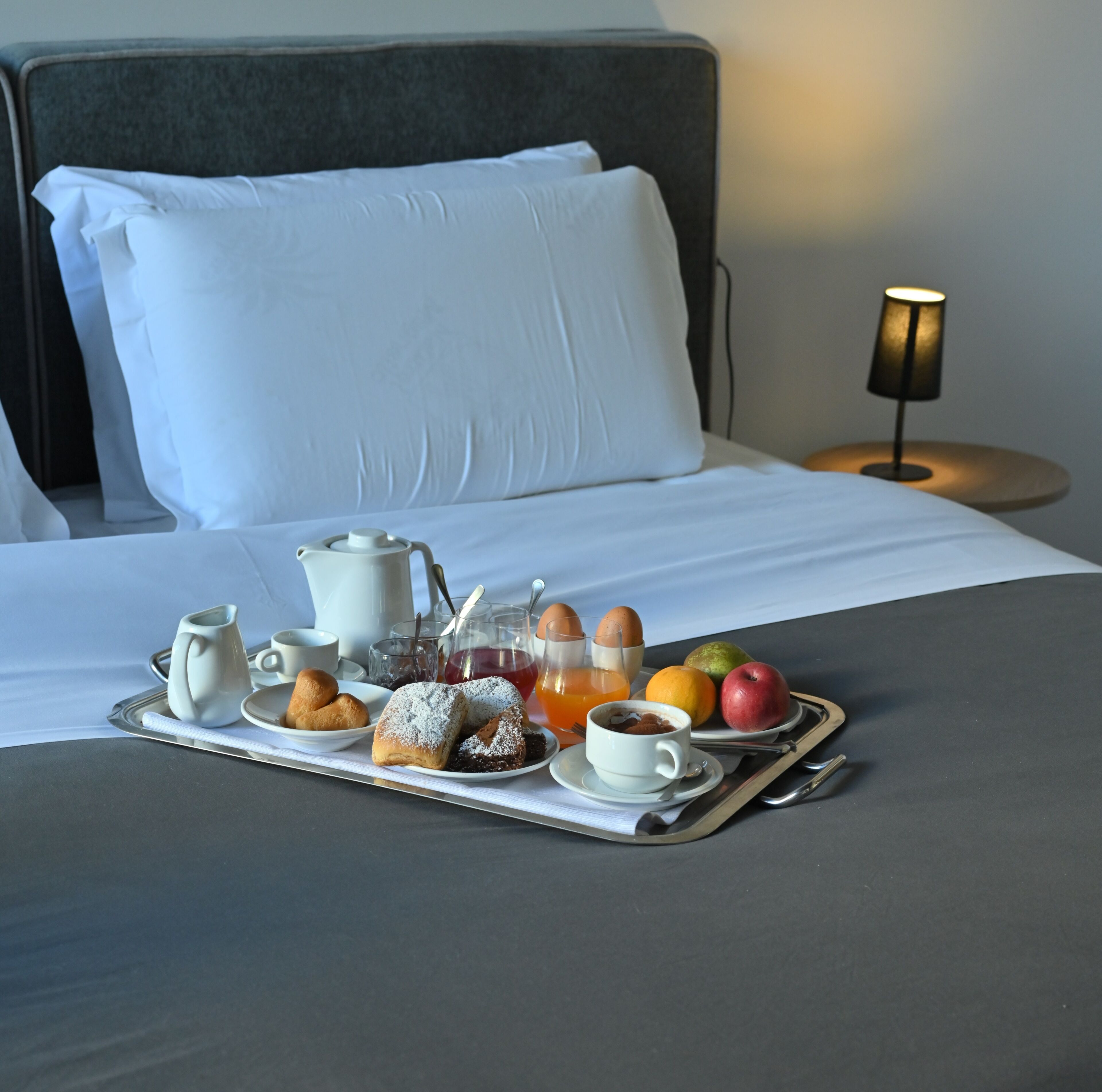 A neatly made breakfast tray on a bed with pillows, a bedside table with a lamp, and a view of the sky.