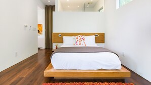 Un dormitorio moderno con cabecera de cama de madera, ropa de cama blanca y vista a un baño a través de una puerta abierta.