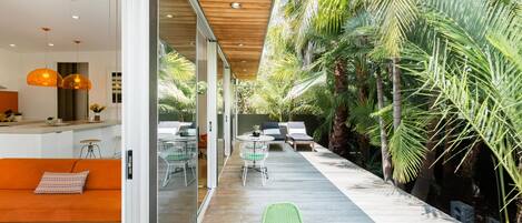 A modern interior with a wooden ceiling, a glass wall, and a wooden deck leading to an outdoor area with palm trees.