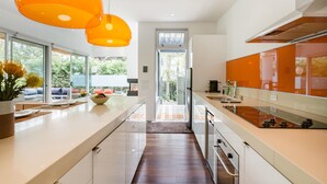 Una cocina moderna con una isla central, ventanas amplias y lámparas colgantes naranjas.
