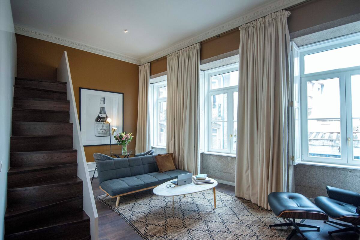 Una sala de estar moderna con una escalera de madera oscura, un sofá gris, una mesa de centro blanca y un sillón de cuero negro.