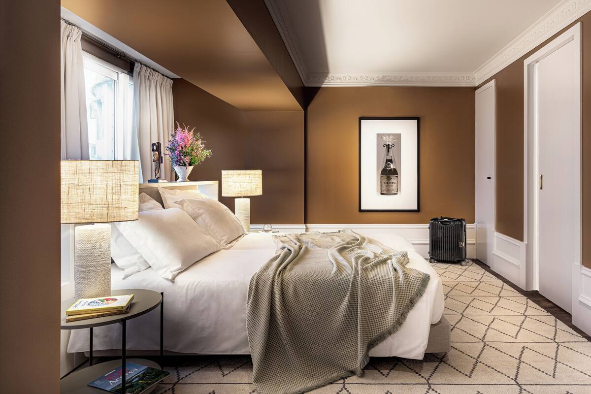 Habitación de hotel con cama, lámparas de noche, una mesita con libros, un cuadro en la pared y una maleta en el suelo.