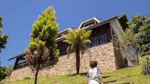 A child walking up a grassy slope towards a stone building with a sloped roof and large windows. - Casa das Montanhas  (Campos do Jordão)