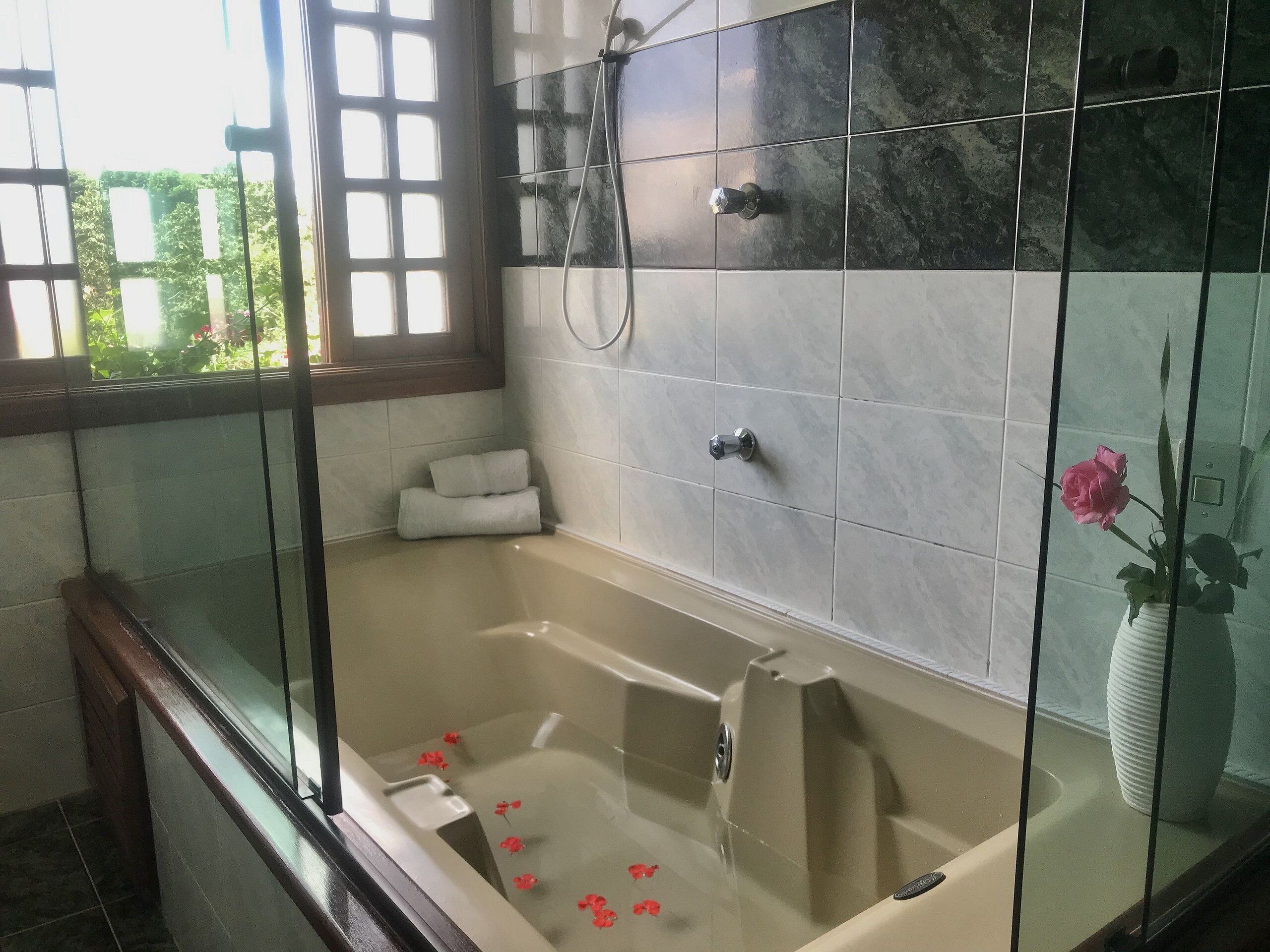 Un baño moderno con ducha envuelta en vidrio, una bañera con pétalos de rosa y una ventana con vistas al verdor.