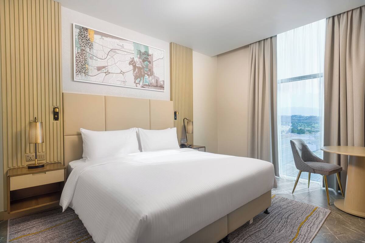 A hotel room with a large bed, a desk, a chair, a map artwork on the wall, and a view of the city through the window.