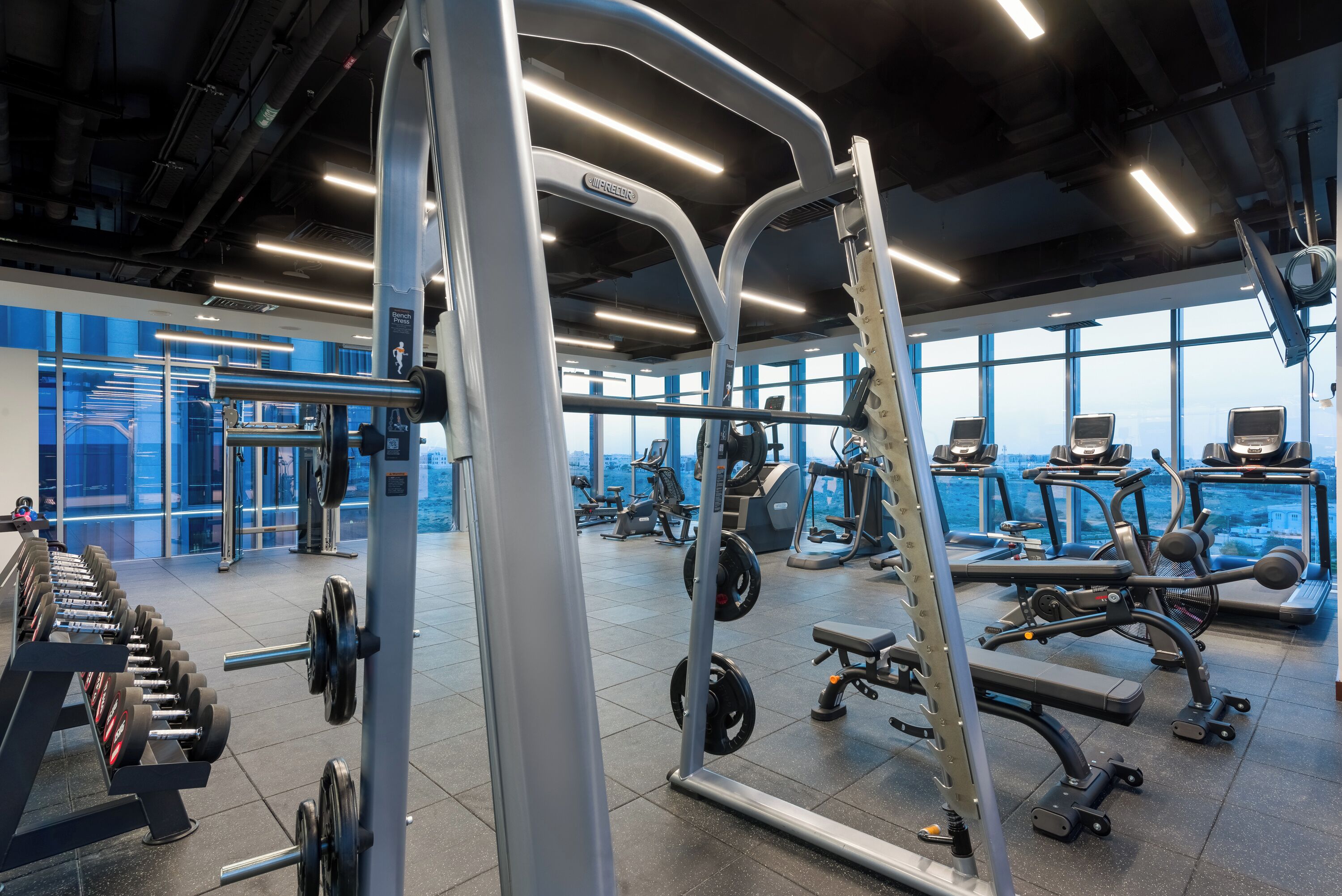 Une salle de sport moderne dotée de divers appareils d’exercice, de grandes fenêtres et d’un plafond haut.