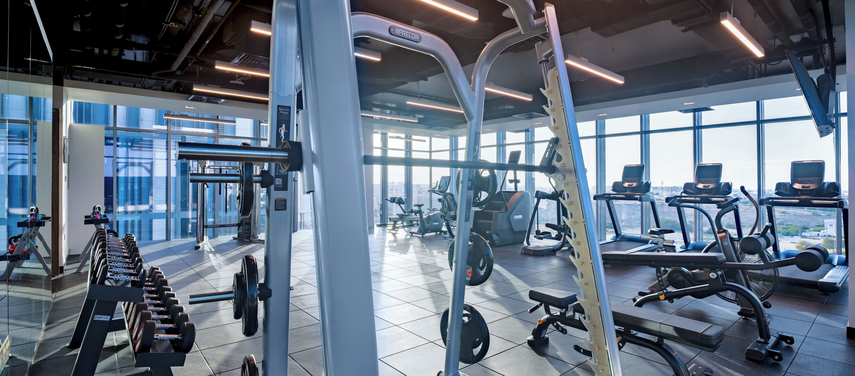 Une salle de sport moderne dotée de divers appareils d’entraînement, de grandes fenêtres et d’un plafond haut.