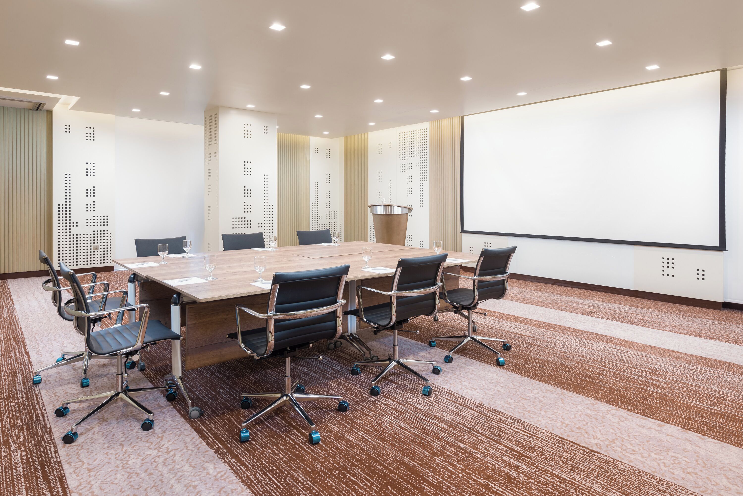 A conference room with a large table, six chairs, a projector screen, and a carpeted floor.