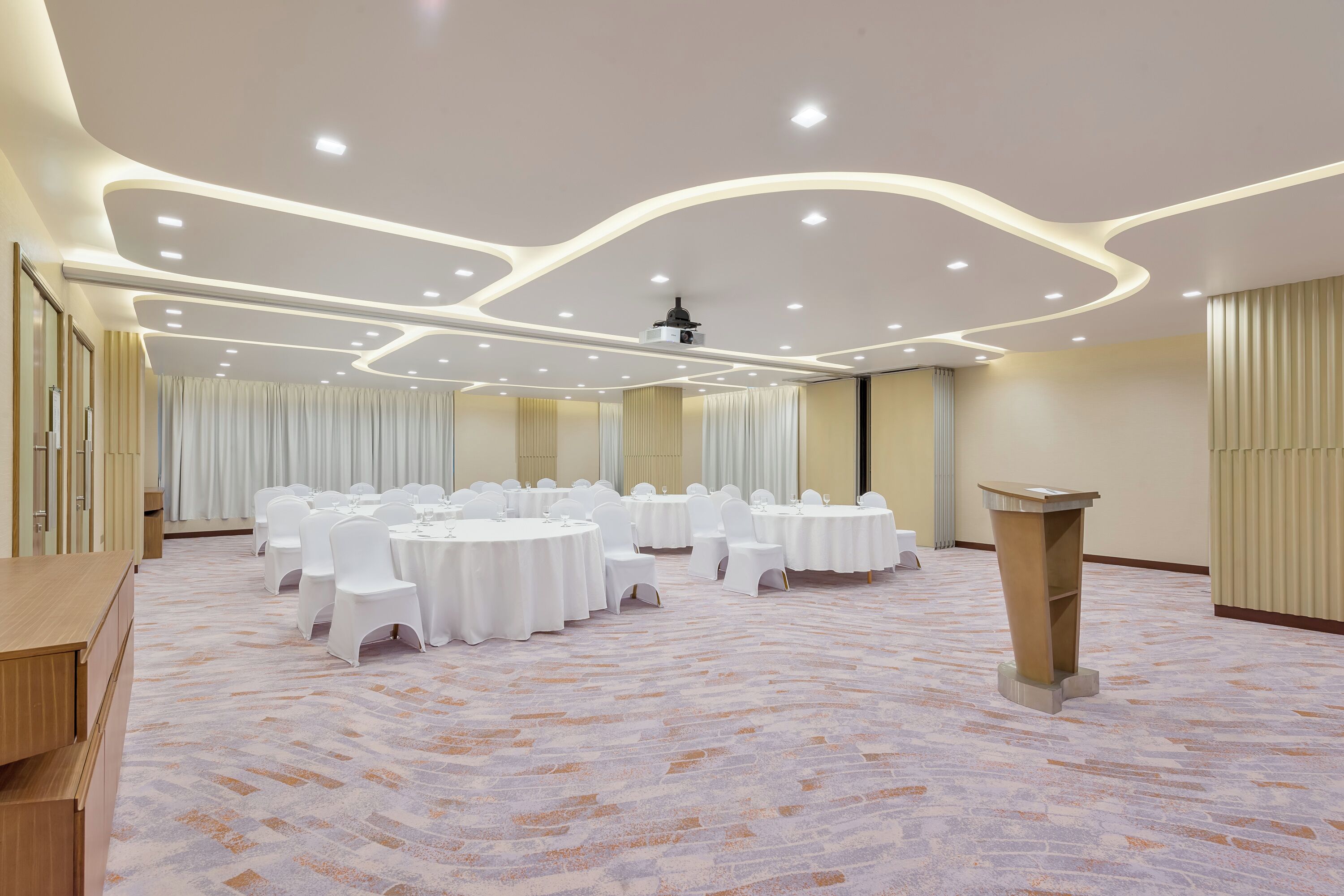 A conference room with multiple round tables, white chairs, a podium, and a carpeted floor.