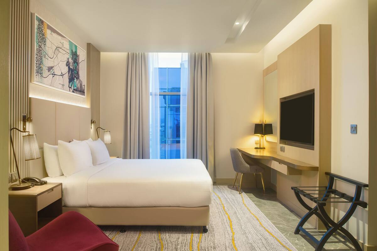 A hotel room with a large bed, a desk, a chair, a TV, and a window with curtains.