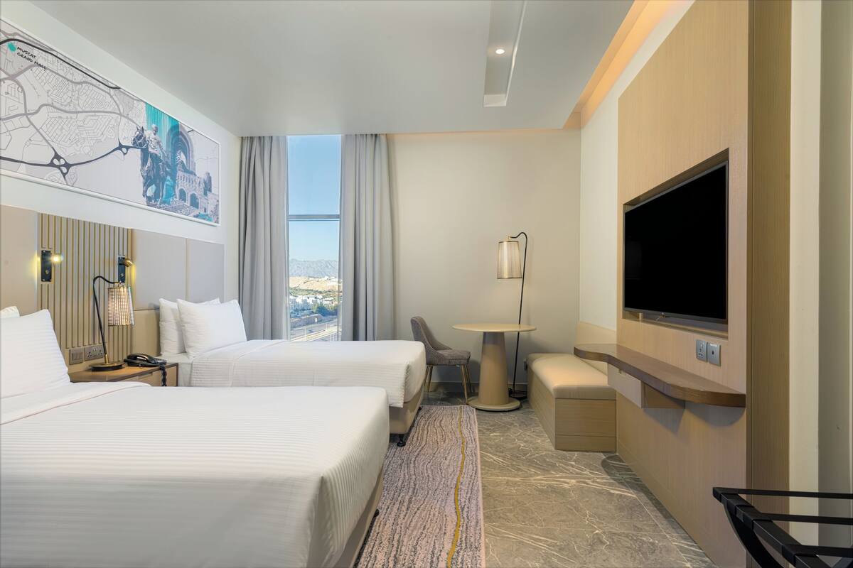 A hotel room with two beds, a desk, a chair, a TV, and a large window with a view.