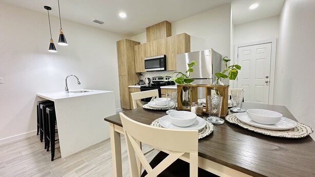 A modern kitchen with a microwave, stove, and refrigerator. There is a dining table set with plates and glasses. - NEW Mcallen Apartament (Pharr)