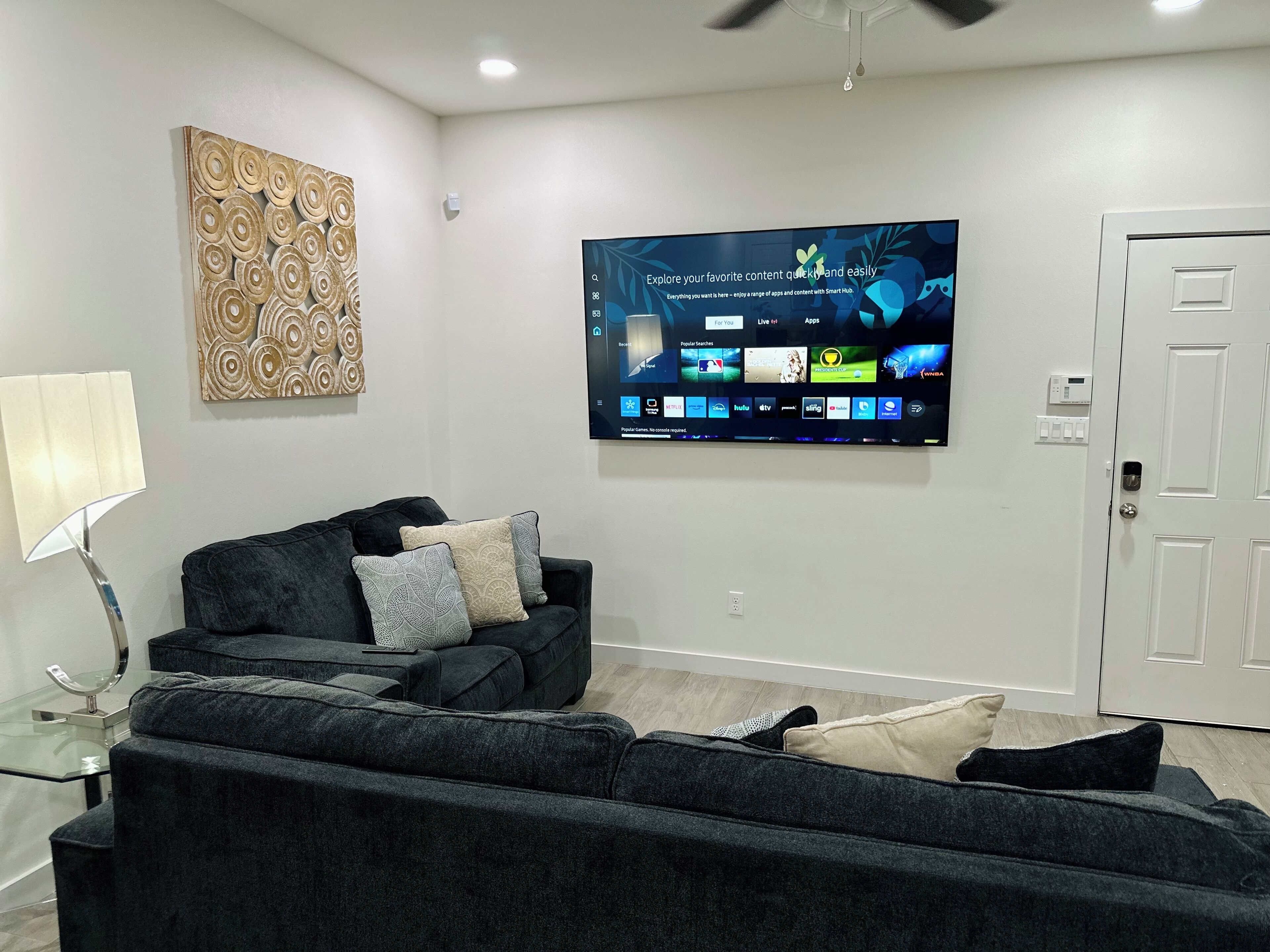 A living room with a flat-screen TV mounted on the wall, a dark-colored sofa with patterned cushions, a glass coffee table, and a modern floor lamp.