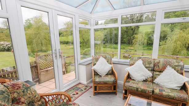 A conservatory with wicker furniture, a glass table, and a view of a garden. - Corran Cottage (Carmarthen)