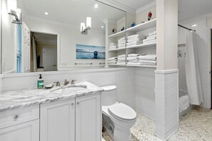 Een moderne badkamer met een witte wastafel, toilet en douchegedeelte. Er zijn planken met opgevouwen handdoeken en een grote spiegel boven de wastafel.