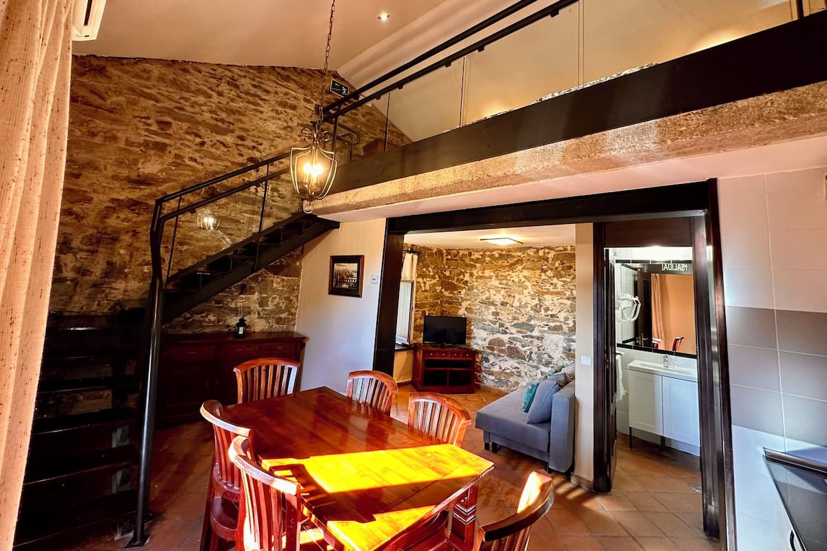 A dining area with a wooden table and chairs, a staircase with a metal railing, and a stone wall.
