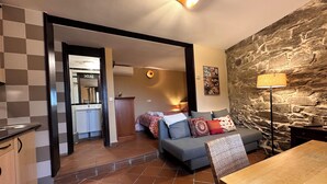 A living room with a stone wall, a sofa, a dining table, and a kitchen area visible through an open doorway. - El Bulín de Patones (Patones)