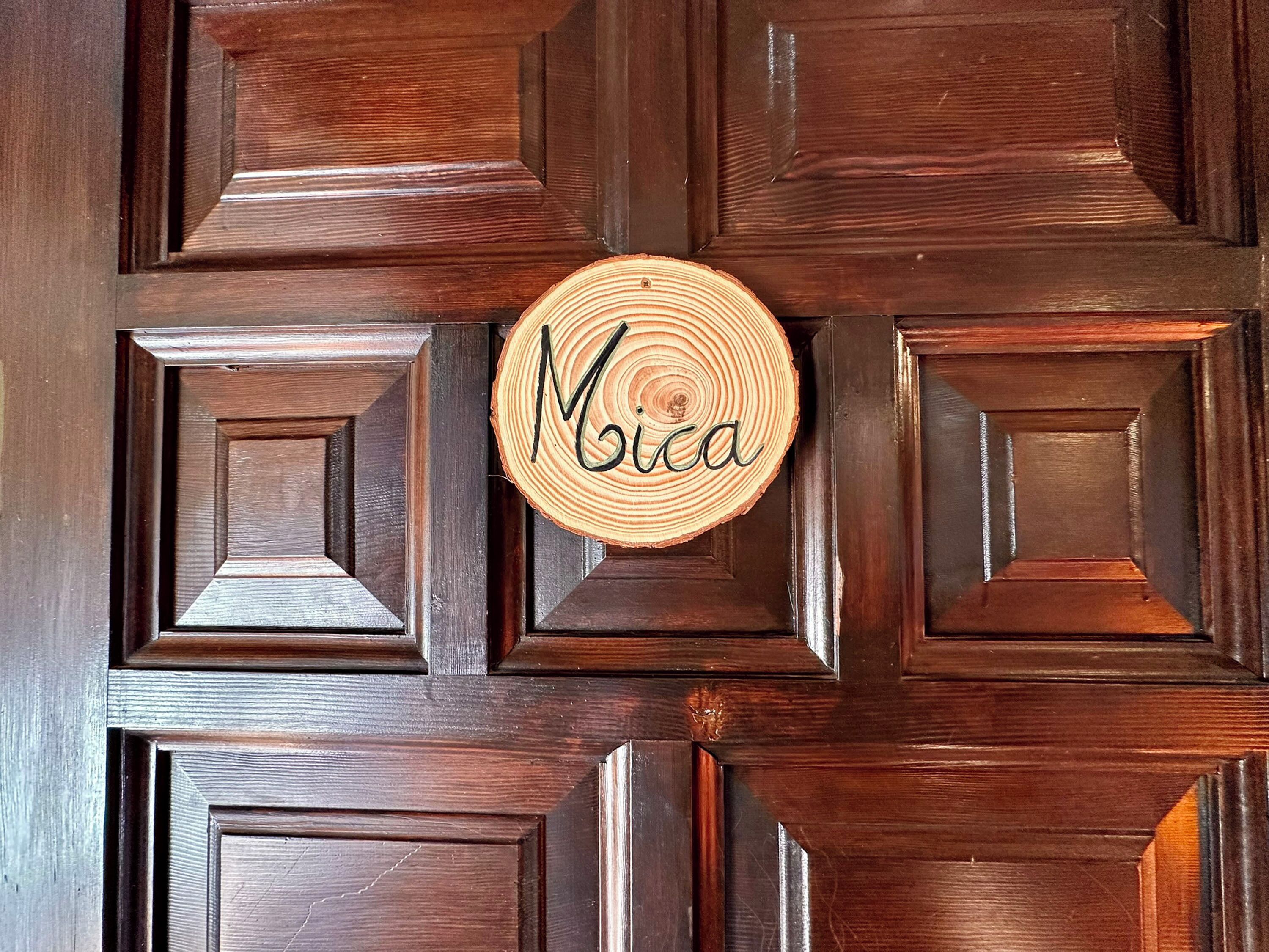 Une porte en bois sur laquelle est fixée une enseigne circulaire en bois, avec le nom « Mica » inscrit au centre.