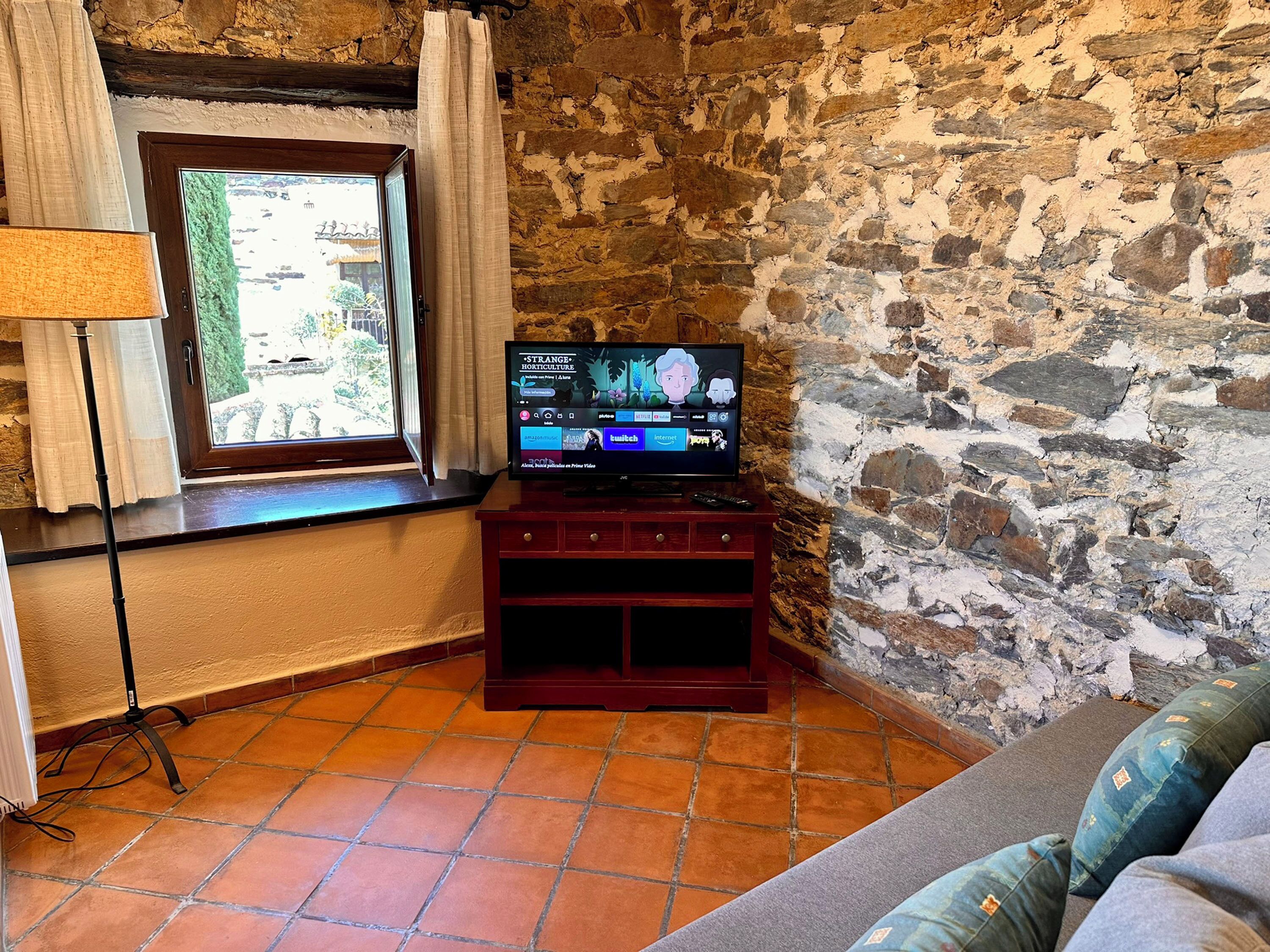 Un salon chaleureux avec un mur en pierre, une fenêtre avec des rideaux, un téléviseur posé sur un meuble en bois et un canapé agrémenté de coussins à motifs.