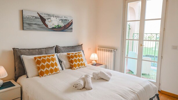 Una cama bien hecha con almohadillas blancas y anaranjadas, una lámpara de noche y un cuadro de un barco en la pared.