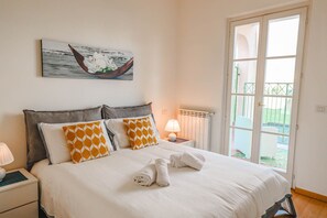 Un letto rifatto con cuscini bianchi e arancioni a motivi decorativi, una lampada da comodino e un quadro raffigurante una barca al muro.