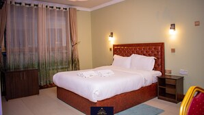Una habitación de hotel con una cama grande, cabecera de madera, mesita de noche, armario y ventana con cortinas.