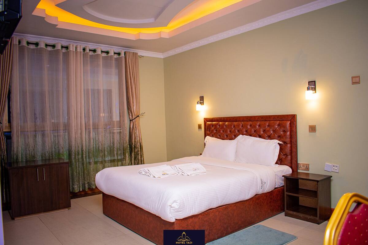 A hotel room with a large bed, a wooden headboard, a nightstand, a wardrobe, and a window with curtains.