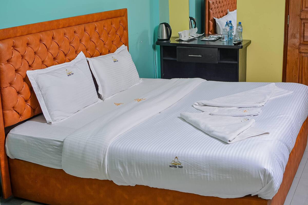 A neatly made bed with white linens and pillows, a wooden headboard, and a bedside table with a kettle, cups, and water bottles.