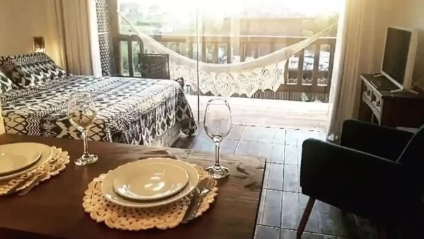 A dining area with a table set for two, a bed with patterned bedding, and a hammock. - Altos da Lagoa-praia do Rosa, Ibiraquera -studio 3 (Imbituba)