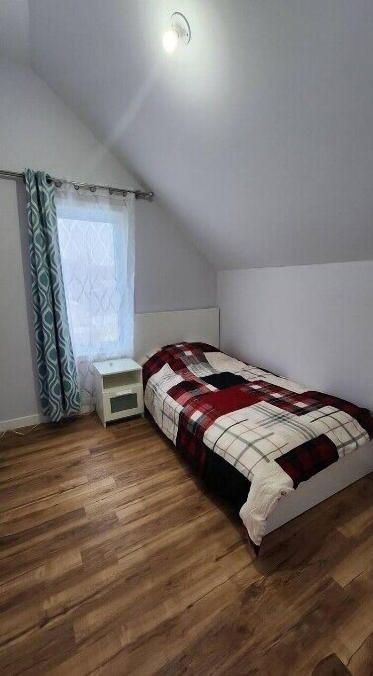 Quarto pequeno com uma cama de solteiro, teto inclinado, mesa de cabeceira, janela com cortinas estampadas e piso de madeira.
