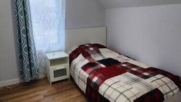 Quarto pequeno com uma cama de solteiro, teto inclinado, mesa de cabeceira, janela com cortinas estampadas e piso de madeira.