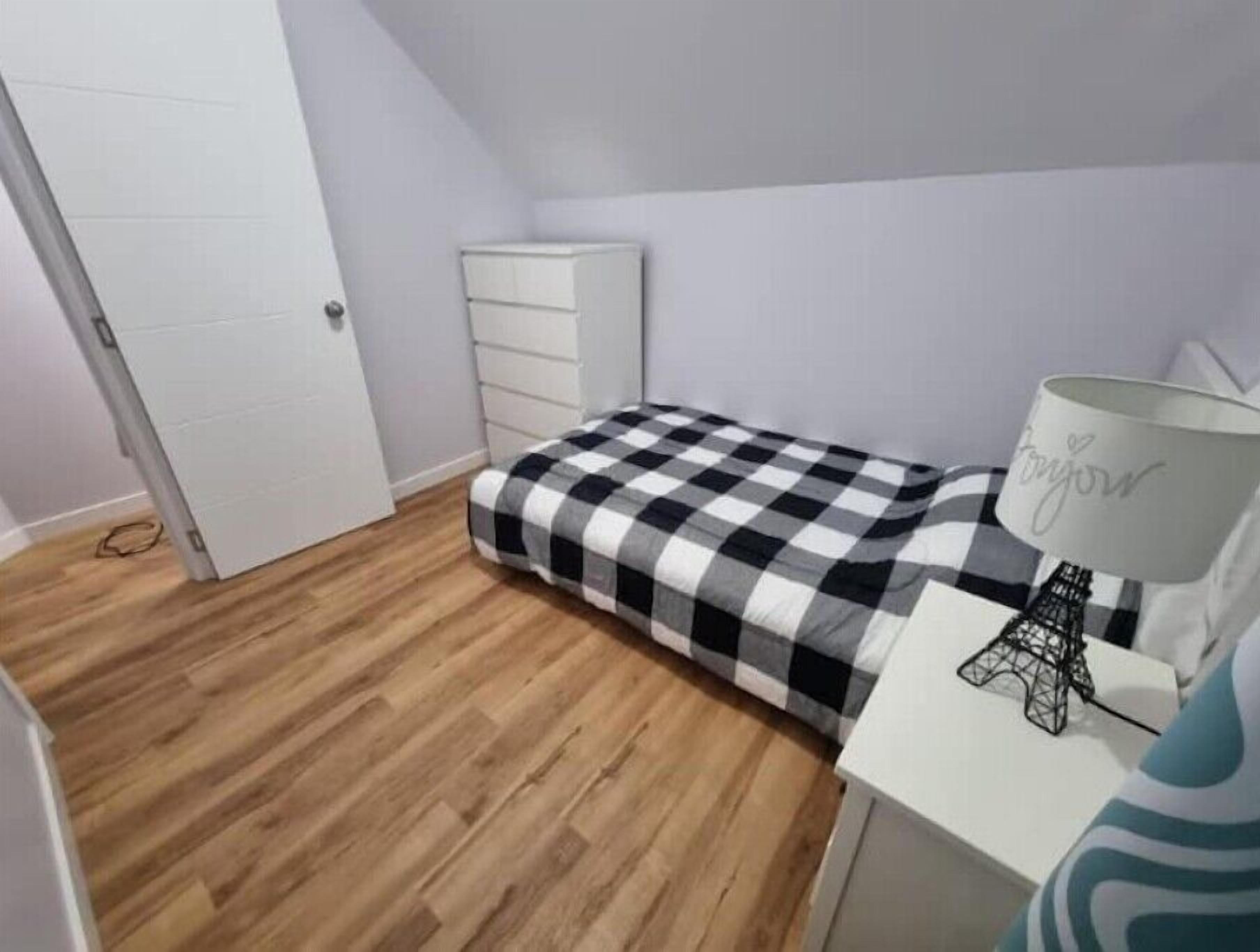 A small bedroom with a checkered bedspread, a white dresser, and a lamp on a bedside table.