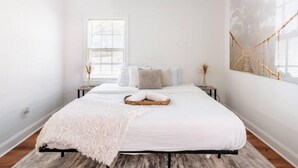 A bedroom with a large bed, a window with blinds, a wall-mounted macrame piece, and a wooden floor. - The Georgia Peach (Forest Park)