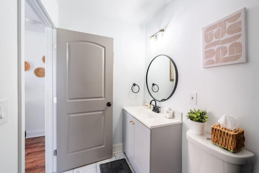 Een badkamer met een witte wastafel, een ronde spiegel, een toilet en een deur naar een andere kamer.