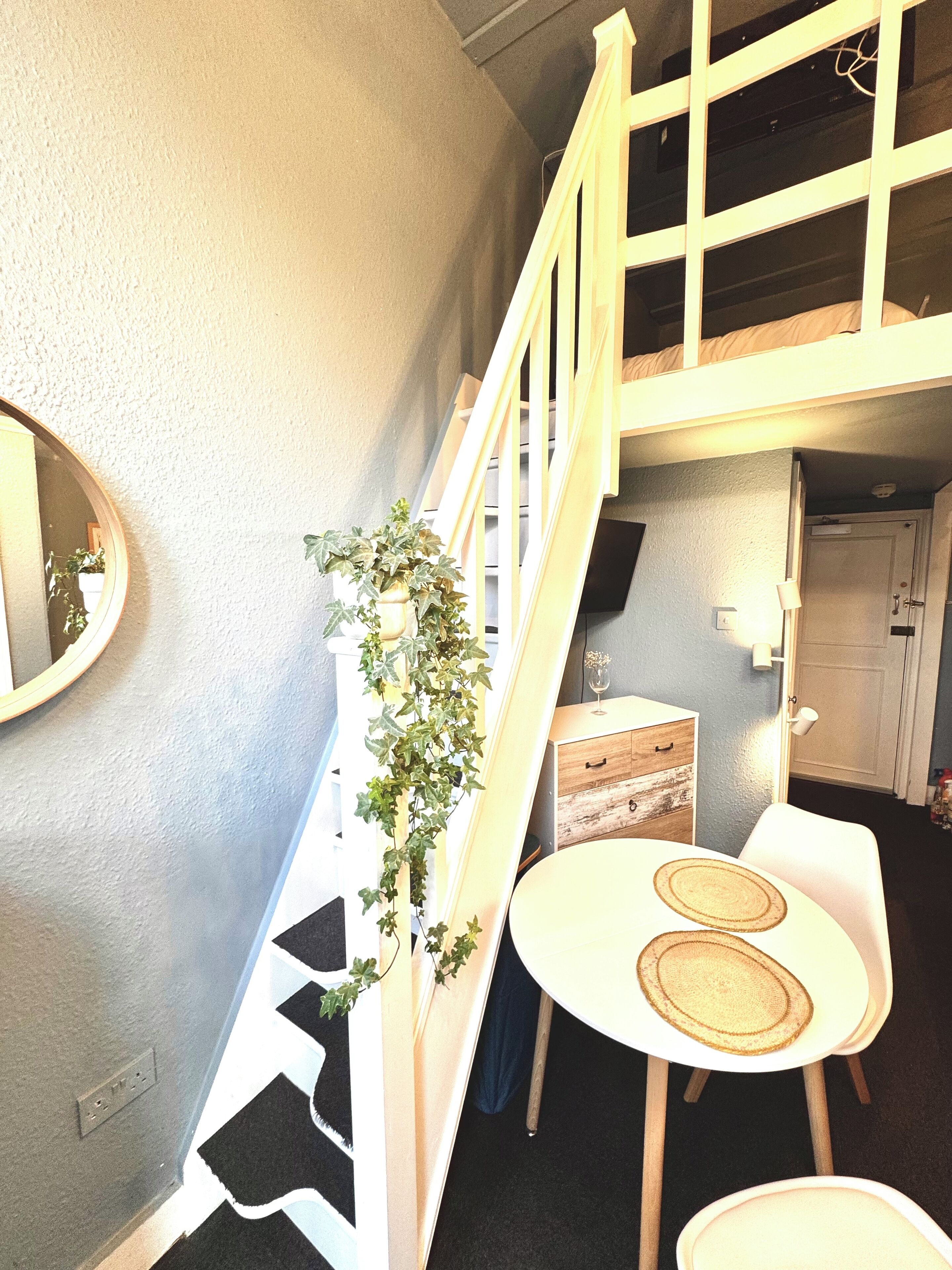 Une pièce avec un escalier, une table à manger avec deux chaises, un petit meuble et une plante en pot.