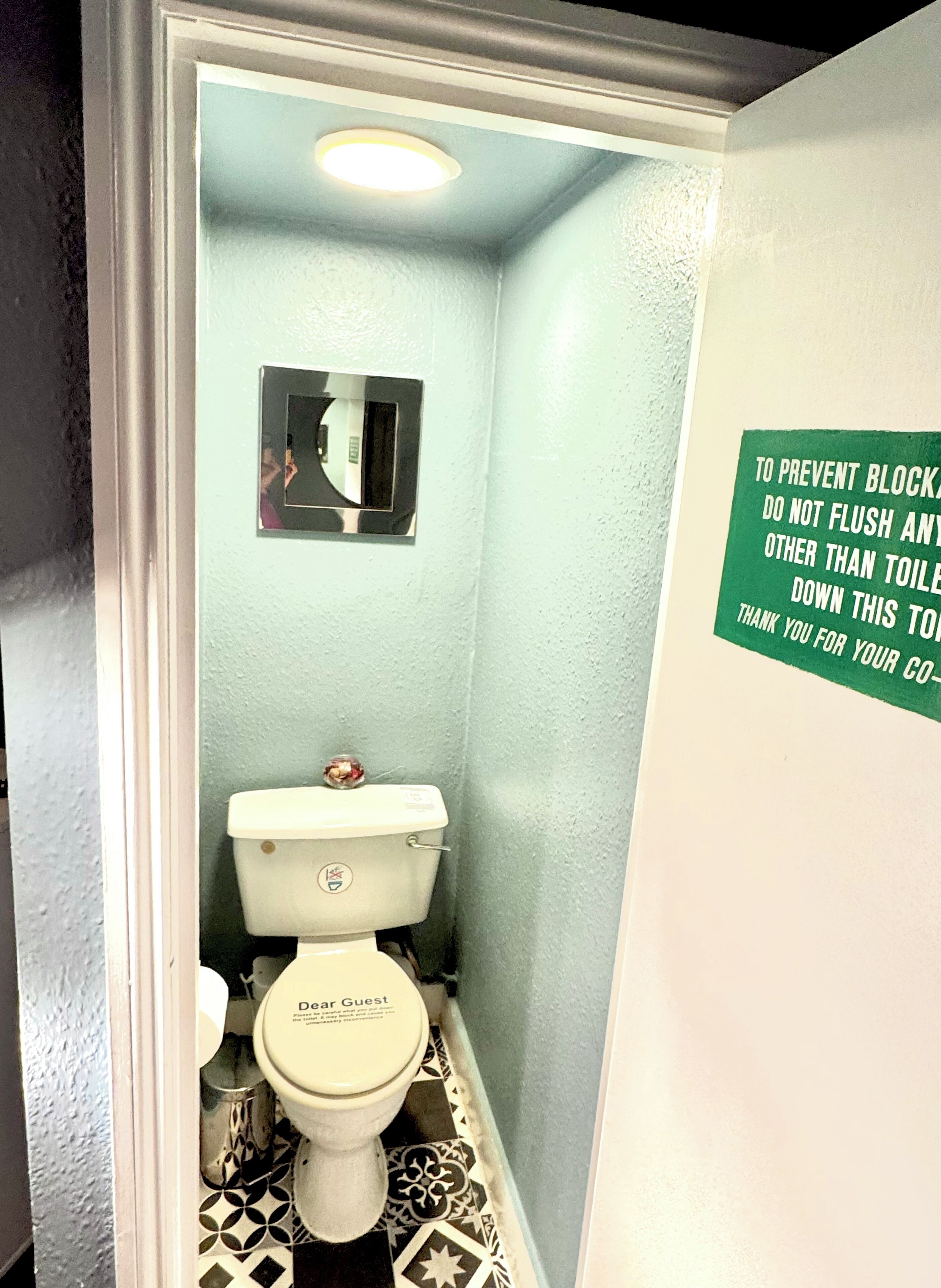 A small, narrow bathroom with a toilet, a mirror, and a sign on the wall.