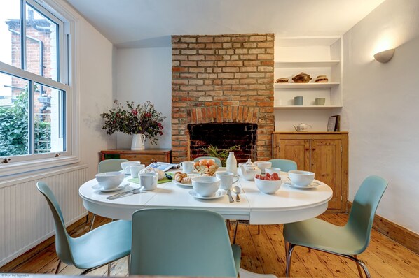 A dining area with a brick fireplace, white walls, and a window overlooking greenery. - Henley Street Townhouse | Oxford Holiday Let | 3 Beds & Sleeps 6 (Oxford)