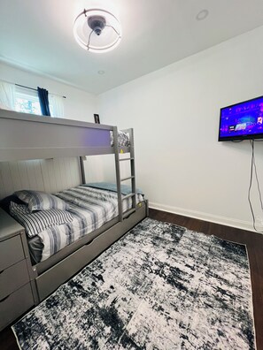 Una cama litera con escalera, un televisor de pantalla plana y un tapete estampado en una habitación con paredes blancas y una lámpara en el techo.
