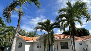 Une maison blanche avec un toit recouvert de tuiles rouges, entourée de palmiers et d’une pelouse verdoyante.