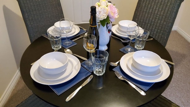 A round dining table set for four with white plates, blue napkins, and clear glasses. A bottle of Prosecco and a vase with flowers are placed in the center. - 8 Guests - 4 Bedrooms - Parking - Garden (Norwich)