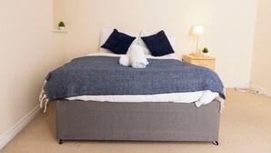A neatly made bed with a grey mattress and blue blanket, a bedside table with a lamp, and a small plant on the nightstand. - 8 Guests - 4 Bedrooms - Parking - Garden (Norwich)