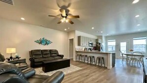 A modern living room with a black leather sofa, a coffee table, and a ceiling fan. There is a kitchen area with a bar and stools, and a dining area with a table and chairs. - New Built - Clean - Comfort (Grand Junction)
