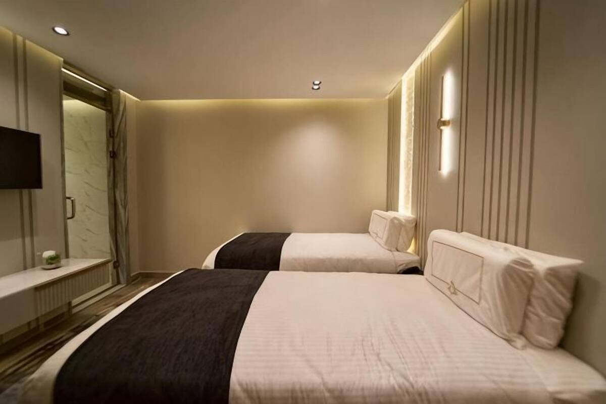 A hotel room with two beds, a TV, a mirror, and a window with vertical blinds.