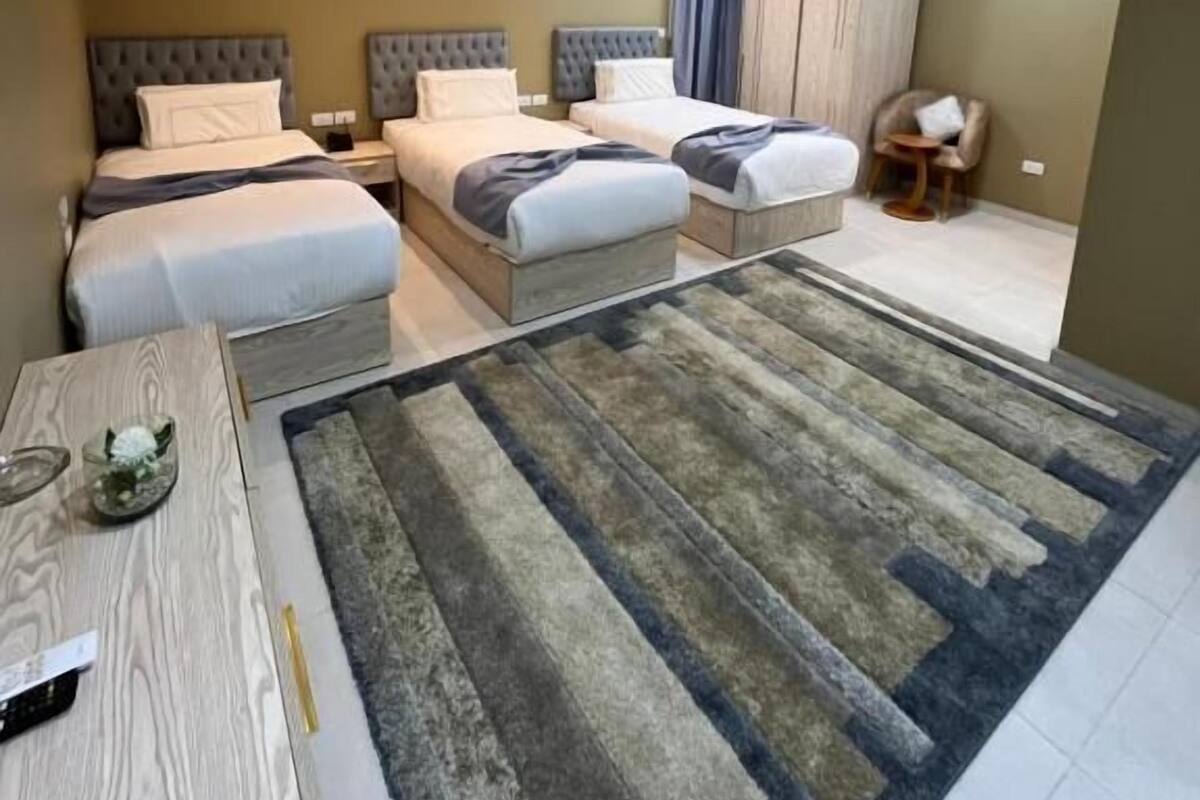 A modern hotel room with two beds, a small table, a chair, and a large rug.
