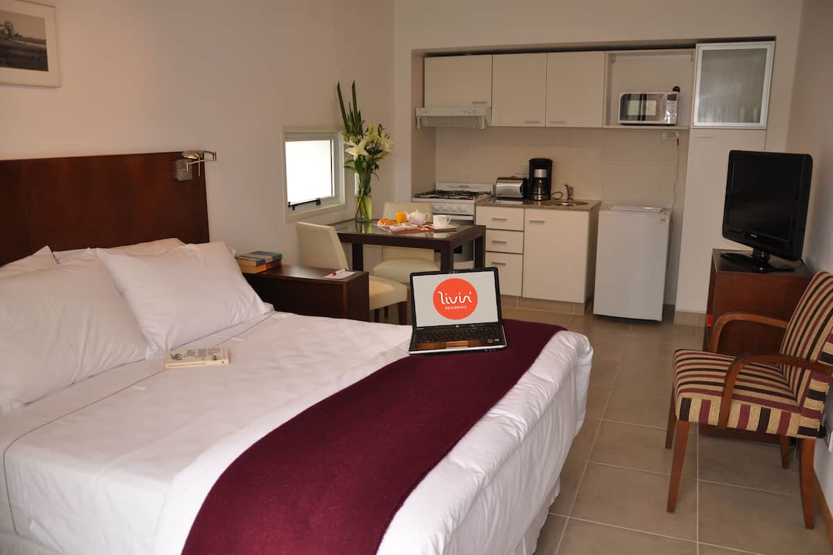 A hotel room with a bed, a desk with a laptop, a chair, a TV, and a kitchenette.
