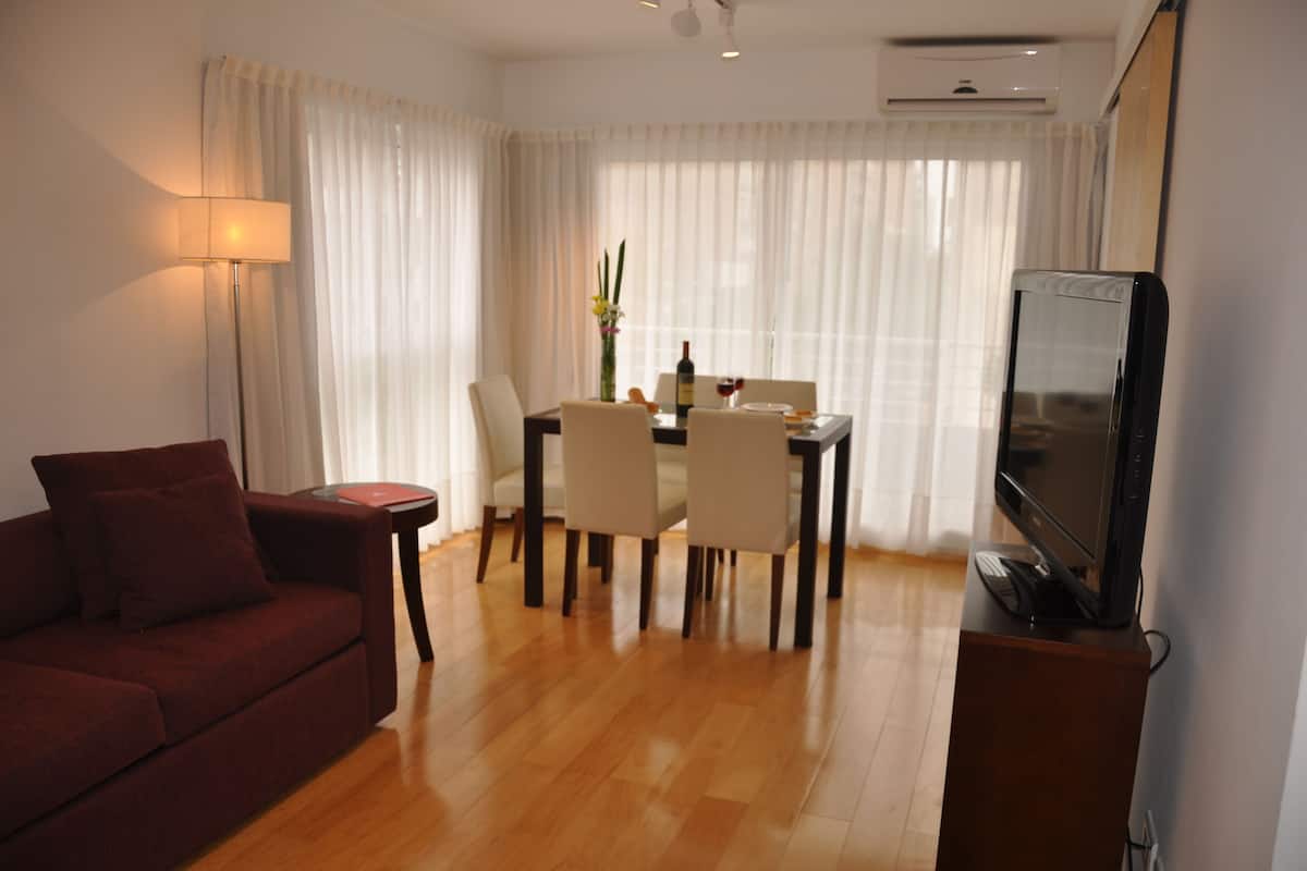 A living room with a sofa, a dining table with chairs, a TV on a stand, and a lamp.